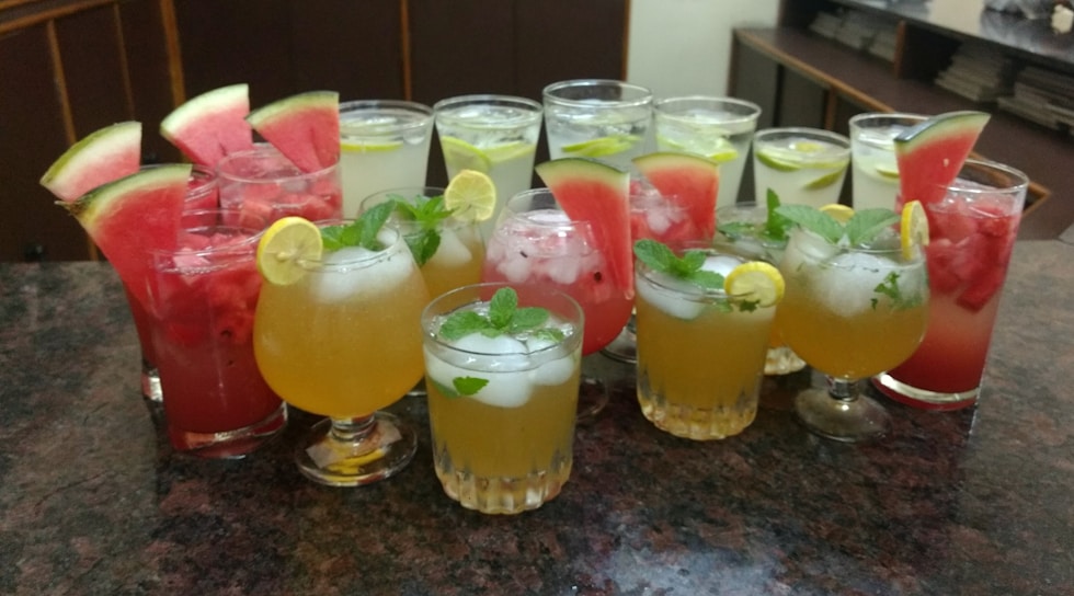 An assortment of colorful beverages is arranged on a granite countertop. The drinks are presented in various types of glassware and garnished with fresh fruits such as watermelon slices, lime wedges, and mint leaves. Ice cubes are visible in some of the glasses, suggesting they are refreshing and cold.