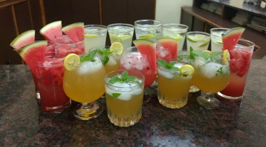 An assortment of colorful beverages is arranged on a granite countertop. The drinks are presented in various types of glassware and garnished with fresh fruits such as watermelon slices, lime wedges, and mint leaves. Ice cubes are visible in some of the glasses, suggesting they are refreshing and cold.