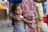 A young child with long dark hair is being held protectively by an adult. The child is wearing a striped shirt and has a concerned expression. The adult is wearing a purple and yellow striped garment with floral patterns, and their arm is wrapped around the child. In the background, there are other blurred figures and a variety of colors.