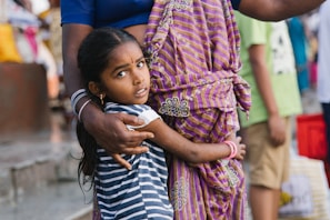 A young child with long dark hair is being held protectively by an adult. The child is wearing a striped shirt and has a concerned expression. The adult is wearing a purple and yellow striped garment with floral patterns, and their arm is wrapped around the child. In the background, there are other blurred figures and a variety of colors.