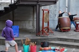 Two children enthusiastically play large traditional Japanese drums on a raised wooden platform, while a child in a purple hoodie and shorts looks on with curiosity. A gong is positioned near the drums, and various percussion instruments are scattered nearby in buckets.