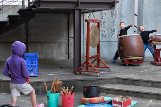 Two children enthusiastically play large traditional Japanese drums on a raised wooden platform, while a child in a purple hoodie and shorts looks on with curiosity. A gong is positioned near the drums, and various percussion instruments are scattered nearby in buckets.