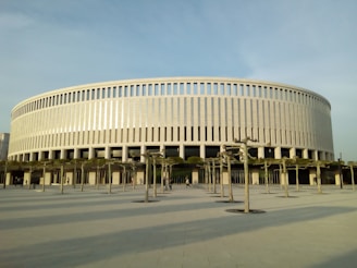 A large, circular building with a modern design featuring vertical columns and a smooth, light-colored facade. In front of the building, there are pruned trees aligned in rows along a paved open space. The sky appears clear, casting soft shadows on the ground.
