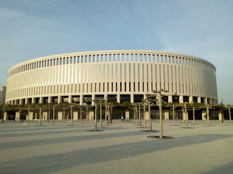 A large, circular building with a modern design featuring vertical columns and a smooth, light-colored facade. In front of the building, there are pruned trees aligned in rows along a paved open space. The sky appears clear, casting soft shadows on the ground.