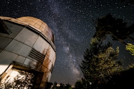 A large, dome-shaped telescope observatory is situated to the left, surrounded by tall trees. The night sky is filled with an expansive view of the Milky Way galaxy, showcasing countless stars. The image has a serene, almost mystical atmosphere.