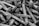 A pile of metallic spikes with rough surfaces, characterized by pointed ends and flat, wide heads. The image is presented in black and white, accentuating the texture of the metal.