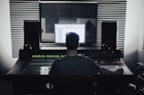 A person is seated in front of an audio mixing console in a recording studio. Large speakers and soundproofing foam panels are visible, with a computer screen displaying audio software.