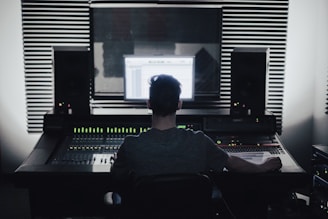A person is seated in front of an audio mixing console in a recording studio. Large speakers and soundproofing foam panels are visible, with a computer screen displaying audio software.