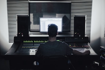 A person is seated in front of an audio mixing console in a recording studio. Large speakers and soundproofing foam panels are visible, with a computer screen displaying audio software.