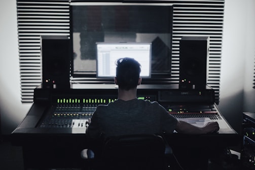 A person is seated in front of an audio mixing console in a recording studio. Large speakers and soundproofing foam panels are visible, with a computer screen displaying audio software.