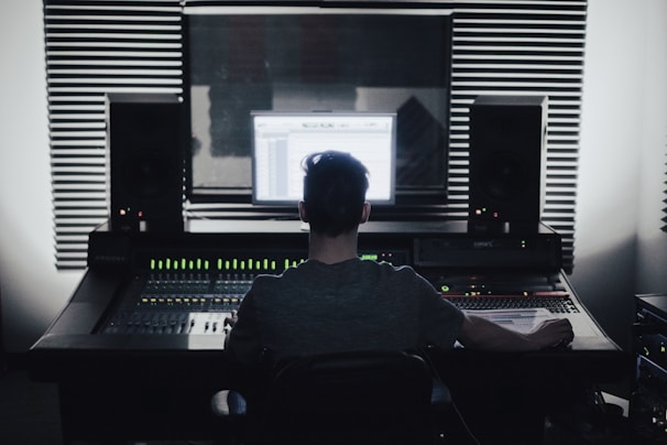 A person is seated in front of an audio mixing console in a recording studio. Large speakers and soundproofing foam panels are visible, with a computer screen displaying audio software.