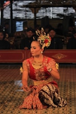A dancer in traditional attire performs with intricate hand gestures. The elaborate costume includes a red and gold top with batik pattern fabric. The dancer wears ornate jewelry and a decorative headpiece adorned with flowers. The background features musicians in traditional garb, adding cultural depth to the scene.