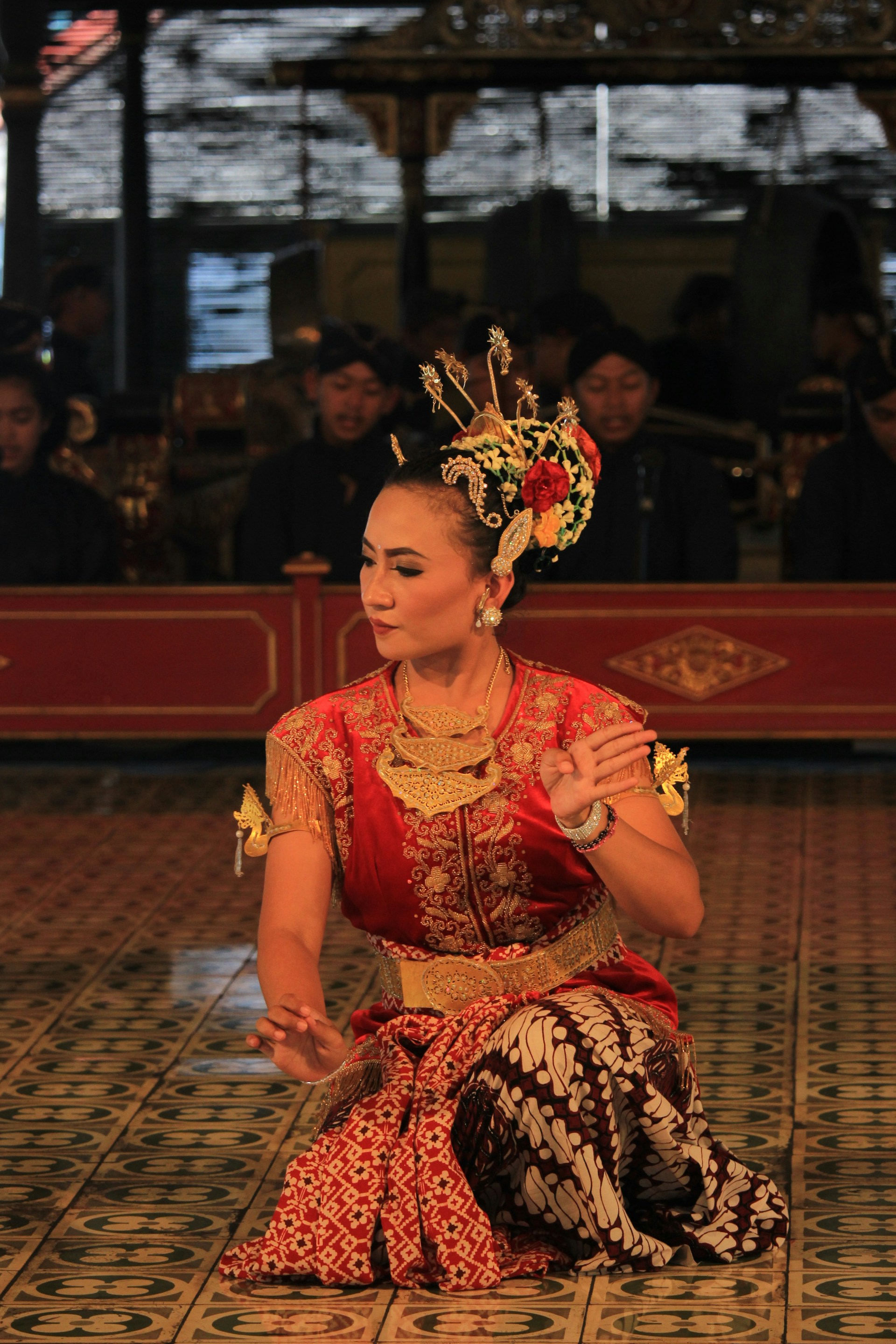 A dancer in traditional attire performs with intricate hand gestures. The elaborate costume includes a red and gold top with batik pattern fabric. The dancer wears ornate jewelry and a decorative headpiece adorned with flowers. The background features musicians in traditional garb, adding cultural depth to the scene.