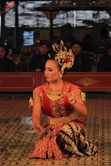 A dancer in traditional attire performs with intricate hand gestures. The elaborate costume includes a red and gold top with batik pattern fabric. The dancer wears ornate jewelry and a decorative headpiece adorned with flowers. The background features musicians in traditional garb, adding cultural depth to the scene.