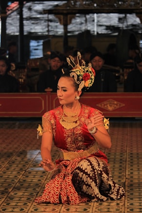 A dancer in traditional attire performs with intricate hand gestures. The elaborate costume includes a red and gold top with batik pattern fabric. The dancer wears ornate jewelry and a decorative headpiece adorned with flowers. The background features musicians in traditional garb, adding cultural depth to the scene.