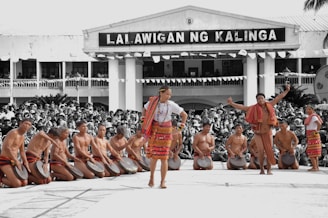 A group of people in traditional attire perform a cultural dance in front of a large crowd. The performers are wearing colorful woven fabrics and are playing handheld drums. The background shows a building with a sign, and the audience is attentively watching the performance.