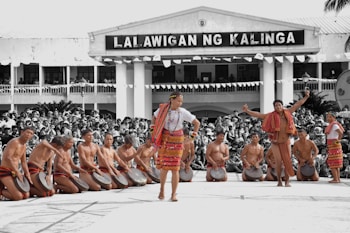A group of people in traditional attire perform a cultural dance in front of a large crowd. The performers are wearing colorful woven fabrics and are playing handheld drums. The background shows a building with a sign, and the audience is attentively watching the performance.