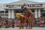 A lively cultural performance is taking place in front of a large crowd. Performers wearing traditional attire are engaged in a synchronized dance. The building in the background has a sign that reads 'LA LALAWIGAN NG KALINGA.' Spectators are actively watching and the atmosphere appears festive.