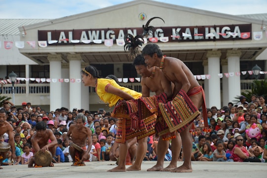 A lively cultural performance is taking place in front of a large crowd. Performers wearing traditional attire are engaged in a synchronized dance. The building in the background has a sign that reads 'LA LALAWIGAN NG KALINGA.' Spectators are actively watching and the atmosphere appears festive.