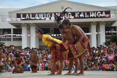 A lively cultural performance is taking place in front of a large crowd. Performers wearing traditional attire are engaged in a synchronized dance. The building in the background has a sign that reads 'LA LALAWIGAN NG KALINGA.' Spectators are actively watching and the atmosphere appears festive.