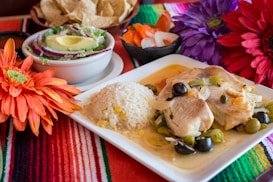 A colorful and vibrant composition featuring a plate of food with rice, fish fillets garnished with olives and vegetables. Accompanied by a bowl of salad with avocado and onion slices. The table is adorned with bold and bright flowers and a colorful striped tablecloth, adding a festive ambiance.