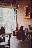 A cozy cafe interior with exposed brick walls and a large letter 'L' illuminated with bulbs. Several people are seated at tables, some engaging in conversation while others are using laptops. There's a large window allowing natural light in, and vintage-style hanging bulbs add a warm glow to the atmosphere.