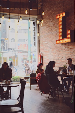 A cozy cafe interior with exposed brick walls and a large letter 'L' illuminated with bulbs. Several people are seated at tables, some engaging in conversation while others are using laptops. There's a large window allowing natural light in, and vintage-style hanging bulbs add a warm glow to the atmosphere.