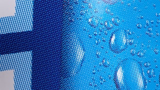 A close-up of a pixelated design with a blue color scheme and water droplets overlaid. The pattern consists of various shades of blue dots forming a gradient from light to dark. The droplets add a realistic, three-dimensional texture to the otherwise flat image.