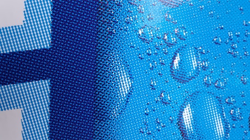 A close-up of a pixelated design with a blue color scheme and water droplets overlaid. The pattern consists of various shades of blue dots forming a gradient from light to dark. The droplets add a realistic, three-dimensional texture to the otherwise flat image.