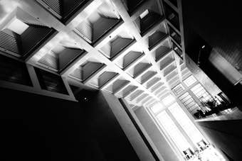 A dramatic, high-contrast black and white photo of a modern architectural interior. The ceiling features geometric recessed panels with a pattern of lines and angles, creating an intriguing play of light and shadow. Tall rectangular windows allow natural light to illuminate the space, enhancing the stark contrast between light and dark areas.