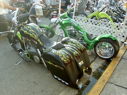 A pair of custom motorcycles with vibrant colors and decorative designs parked on a street. One motorcycle features a bold green color scheme with chrome wheels, while the other is adorned with flame patterns in black and yellow. People are visible in the background, mingling and observing the bikes.