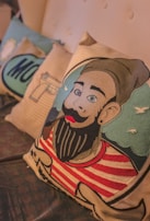 A decorative pillow featuring an illustration of a character with a beanie and a red and white striped shirt, holding a pipe in their mouth. The background includes a light blue sky with small white clouds, and other pillows with various designs are partially visible in the background.