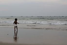 A young child wearing a diaper stands on a beach, facing the ocean. The waves gently approach the shore, and a line of small boats can be seen on the horizon under a cloudy sky.