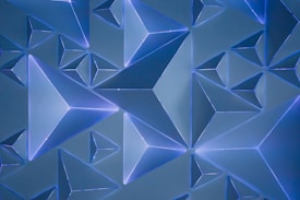 A geometric pattern of three-dimensional triangular prisms with glowing edges, arranged against a blue background. The prisms are predominantly blue with lighter and darker shades, creating a sense of depth and movement.