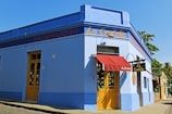 A vibrant corner of a blue building with a red canopy over the entrance. The facade has yellow doors and signage with the words 'La Carlota' and 'Almacen'. Large windows display various items, and a tree and blue sky can be seen in the background.