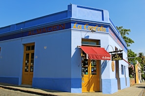 A vibrant corner of a blue building with a red canopy over the entrance. The facade has yellow doors and signage with the words 'La Carlota' and 'Almacen'. Large windows display various items, and a tree and blue sky can be seen in the background.