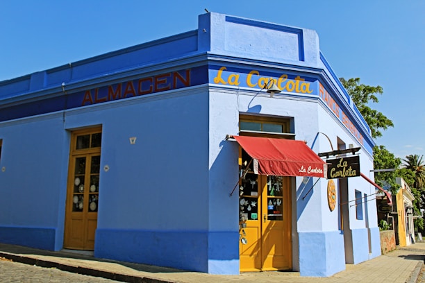 A vibrant corner of a blue building with a red canopy over the entrance. The facade has yellow doors and signage with the words 'La Carlota' and 'Almacen'. Large windows display various items, and a tree and blue sky can be seen in the background.