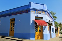 A vibrant corner of a blue building with a red canopy over the entrance. The facade has yellow doors and signage with the words 'La Carlota' and 'Almacen'. Large windows display various items, and a tree and blue sky can be seen in the background.