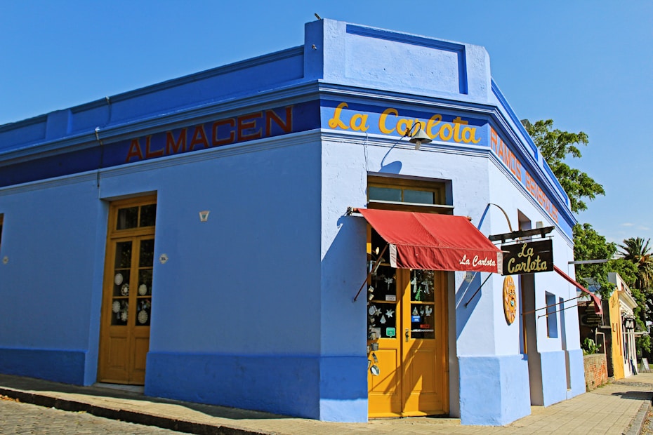 A vibrant corner of a blue building with a red canopy over the entrance. The facade has yellow doors and signage with the words 'La Carlota' and 'Almacen'. Large windows display various items, and a tree and blue sky can be seen in the background.