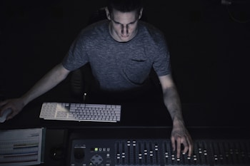 A person is seated at a control panel with a keyboard and a mixing console, concentrating intently, likely engaged in audio or video production. The room is dimly lit, highlighting the person's focused expression and the glowing equipment.