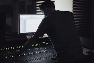 A person is working at an audio mixing console in a dimly lit recording studio. The backlit screen shows audio tracks, and there are soundproofing panels on the walls.