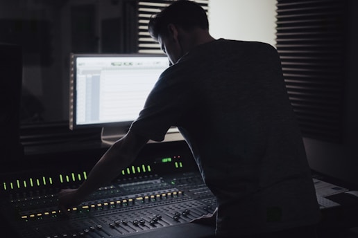 A person is working at an audio mixing console in a dimly lit recording studio. The backlit screen shows audio tracks, and there are soundproofing panels on the walls.