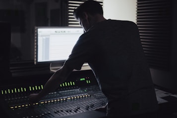 A person is working at an audio mixing console in a dimly lit recording studio. The backlit screen shows audio tracks, and there are soundproofing panels on the walls.