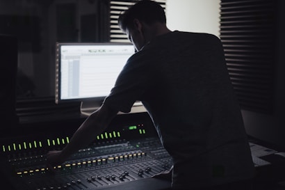 A person is working at an audio mixing console in a dimly lit recording studio. The backlit screen shows audio tracks, and there are soundproofing panels on the walls.