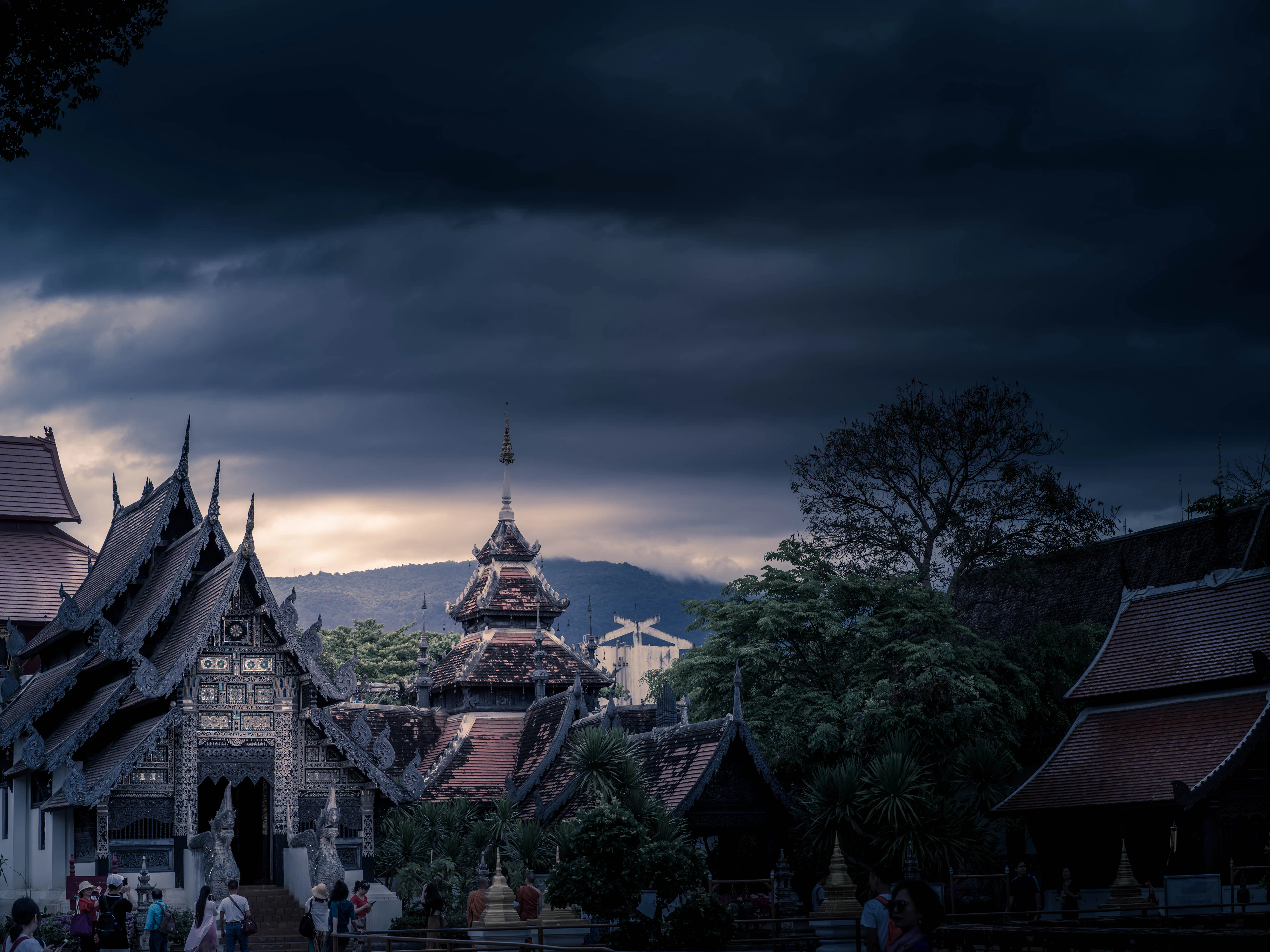 Heavy storm clouds gathering at dusk over the tiered roofs and gilded spire of a Lanna temple compound in Chiang Mai, with hills on the horizon