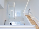 An interior space with a modern, minimalist design characterized by clean lines and open areas. A prominent wooden staircase with built-in lighting leads to the upper floors, surrounded by white walls and large windows that allow natural light to flood the space.