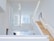 An interior space with a modern, minimalist design characterized by clean lines and open areas. A prominent wooden staircase with built-in lighting leads to the upper floors, surrounded by white walls and large windows that allow natural light to flood the space.