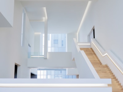 An interior space with a modern, minimalist design characterized by clean lines and open areas. A prominent wooden staircase with built-in lighting leads to the upper floors, surrounded by white walls and large windows that allow natural light to flood the space.
