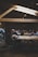 A dimly lit coffee shop features wooden beams on the ceiling and large communal tables. People are sitting on stools, engaging in conversations, working on laptops, and sipping drinks. The atmosphere suggests a casual, modern setting with a focus on both work and social interaction.