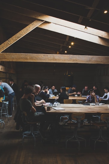 A dimly lit coffee shop features wooden beams on the ceiling and large communal tables. People are sitting on stools, engaging in conversations, working on laptops, and sipping drinks. The atmosphere suggests a casual, modern setting with a focus on both work and social interaction.
